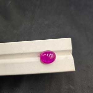 Puede incluir: Una vibrante gema magenta, de forma ovalada, descansa sobre una superficie blanca. La piedra pulida refleja la luz, mostrando su forma suave y redondeada. La gema se encuentra sobre un fondo oscuro, resaltando su color y claridad. Adecuada para la fabricación de joyas.