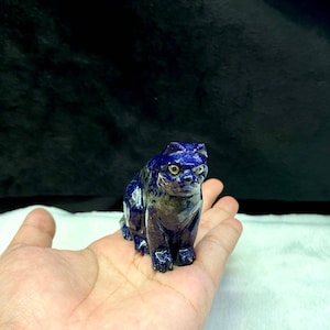 Op de afbeelding: Een klein, gesneden kattenbeeldje gemaakt van blauwe lapis lazuli edelsteen. De kat zit met zijn hoofd lichtjes gekanteld en heeft gouden ogen.
