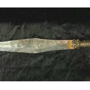 May include: A long, silver-colored spear with a brown wooden handle. The spear has a pointed tip and a decorative design on the handle.