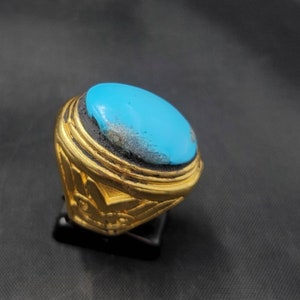 Könnte beinhalten: Ein goldener Ring mit einem großen ovalen Türkis-Stein, der in der Mitte eingesetzt ist. Der Ring hat komplizierte Details um den Stein und auf der Band.