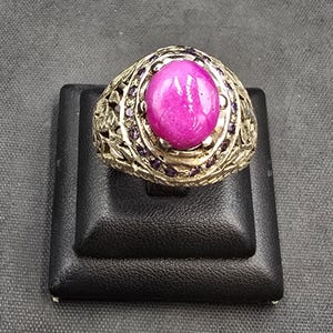 Puede incluir: Un anillo de oro con una gran piedra preciosa ovalada de color rosa rodeada de piedras preciosas violetas más pequeñas.
