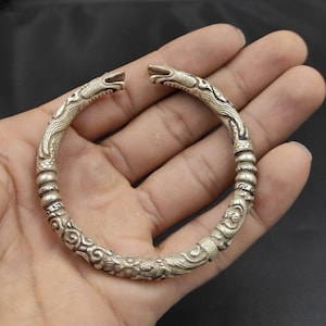 May include: A silver bracelet with a dragon design. The bracelet is open and has a detailed design of a dragon with scales and claws.