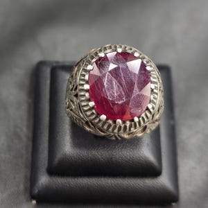 May include: A silver ring with an ornate design and a large, round, red ruby gemstone.