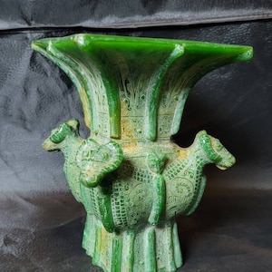 Puede incluir: Un jarrón de jade verde con intrincados tallados y tres cabezas de carnero en los lados. El jarrón tiene una base cuadrada y una parte superior ancha y plana.