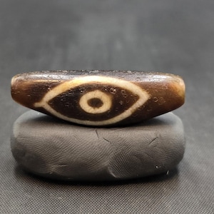 Puede incluir: Una cuenta cil&iacute;ndrica de piedra marr&oacute;n con un patr&oacute;n central en forma de ojo blanco y una pupila circular de color marr&oacute;n oscuro en el centro. La cuenta tiene una superficie texturizada y desgastada, colocada horizontalmente sobre una base gris.