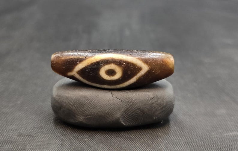 Puede incluir: Una cuenta cil&iacute;ndrica de piedra marr&oacute;n con un patr&oacute;n central en forma de ojo blanco y una pupila circular de color marr&oacute;n oscuro en el centro. La cuenta tiene una superficie texturizada y desgastada, colocada horizontalmente sobre una base gris.