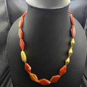 May include: A necklace with alternating red and gold-colored beads. The red beads are diamond-shaped, and the gold beads are cylindrical. The necklace is displayed on a black mannequin.