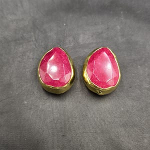 May include: A pair of gold earrings with pink gemstone drops. The earrings are teardrop shaped and have a faceted design.