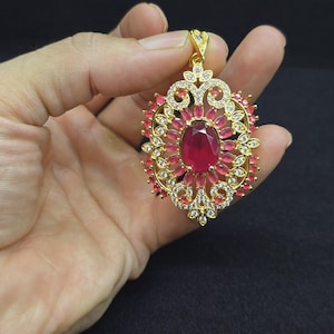 May include: A gold-toned pendant featuring a large central oval-cut red gemstone surrounded by smaller oval-cut red stones and clear white accent stones. The metal frame has an intricate filigree design with a triangular bail at the top.