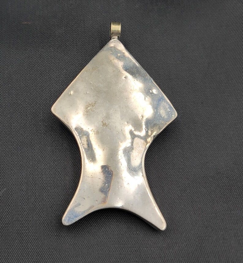 May include: A silver pendant in the shape of a stylized fish with a loop at the top for attaching a chain.