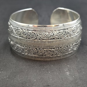 May include: A wide, silver-colored cuff bracelet with intricate floral and scrollwork patterns. The open-ended design allows for easy wear. The bracelet's surface is detailed with multiple bands of embossed designs, creating a textured appearance.
