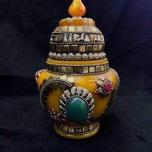 Puede incluir: Un jarrón decorativo de ámbar amarillo con una tapa de oro y plata. El jarrón está decorado con detalles dorados y plateados, y una piedra verde.