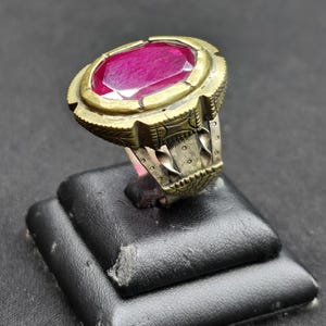 Puede incluir: Un anillo con una gran piedra preciosa de color rosa intenso y corte ovalado, engastada en un bisel de metal con tono de latón envejecido y texturizado. La banda es de metal plateado con grabados, alambres trenzados y perforaciones.