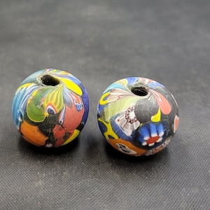 Puede incluir: Dos cuentas de vidrio coloridas con un patrón de remolino en tonos de azul, rojo, amarillo, verde y negro. Las cuentas tienen un agujero en el centro para enhebrar.