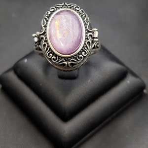 Op de afbeelding: Een zilveren ring met een medaillon met een ovale, paarse steen. De ring heeft een gedetailleerd, versierd ontwerp.