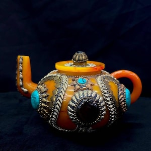 May include: A yellow teapot with intricate silver and gold detailing, adorned with turquoise and black gemstones. The teapot has a rounded body and a curved spout.