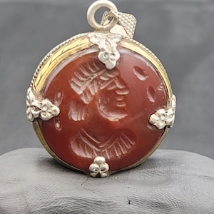 Peut inclure: Un pendentif en argent avec une pierre de cornaline rouge ornée d'un motif sculpté. La pierre est entourée d'un cadre en argent avec des accents dorés.
