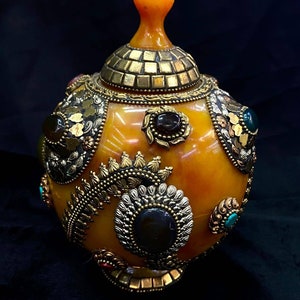 Op de afbeelding: Een ronde, amberkleurige decoratieve container met een gouden deksel en ingewikkeld metaalwerk. De container is versierd met verschillende edelstenen, waaronder turquoise, groen en zwart.