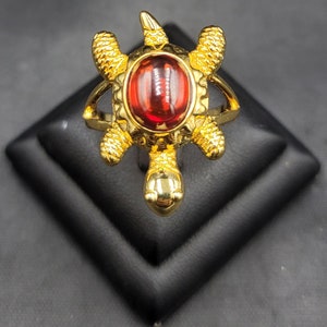 May include: A gold-colored turtle ring with a red oval-shaped stone in the center. The ring features detailed turtle design elements, including the head, legs, and shell. The ring is displayed on a black surface.