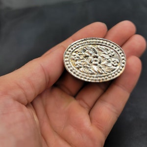 Könnte beinhalten: Eine ovale Brosche aus Silber mit einem detaillierten Blumendesign. Die Brosche hat eine erhabene, strukturierte Oberfläche und einen glatten, polierten Rand.