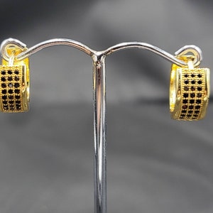 Könnte beinhalten: Ein Paar goldene Creolen mit schwarzen Zirkonia-Steinen. Die Ohrringe befinden sich auf einem silbernen Ständer.