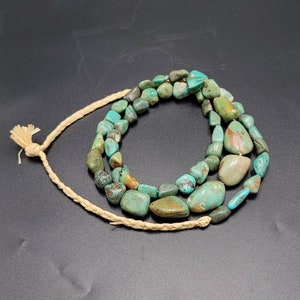 May include: A turquoise gemstone necklace with a natural, irregular shape. The necklace is made of multiple strands of turquoise beads and is strung on a natural fiber cord.