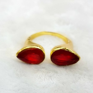May include: A gold-colored ring with two teardrop-shaped, bright red gemstones. The ring has an open design, with the gemstones facing outward. The ring is set against a white background.