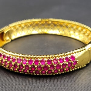 Puede incluir: Un brazalete de oro con una fila de piedras preciosas rosas alrededor del borde.