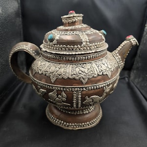 May include: An ornate, antique teapot with a copper body and silver-colored embellishments. The teapot features intricate designs, including floral and animal motifs, and is adorned with turquoise and red gemstones. The handle and spout are also detailed.