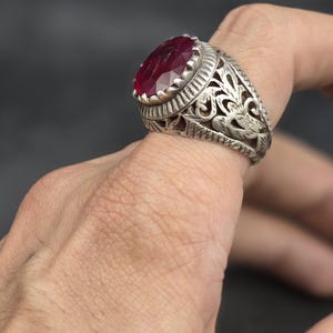 May include: A silver ring with an ornate design featuring a large, oval-shaped, red gemstone.