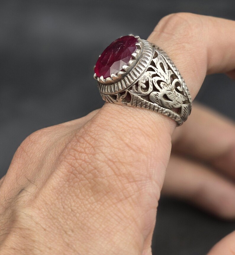 May include: A silver ring with an ornate design featuring a large, oval-shaped, red gemstone.