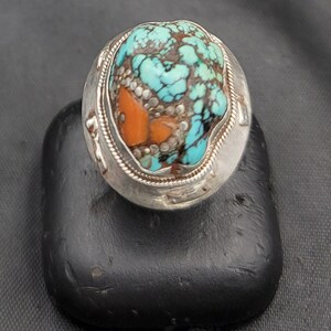 Könnte beinhalten: Ein silberner Ring mit einem großen Türkis- und Orangenstein, der in der Mitte eingesetzt ist. Der Stein ist von einem silbernen Band mit einem dekorativen Design umgeben.