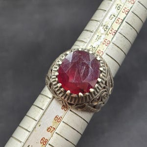 May include: A silver ring with an ornate design and a large, red gemstone. The ring is being measured with a ruler, and the measurement is 67.