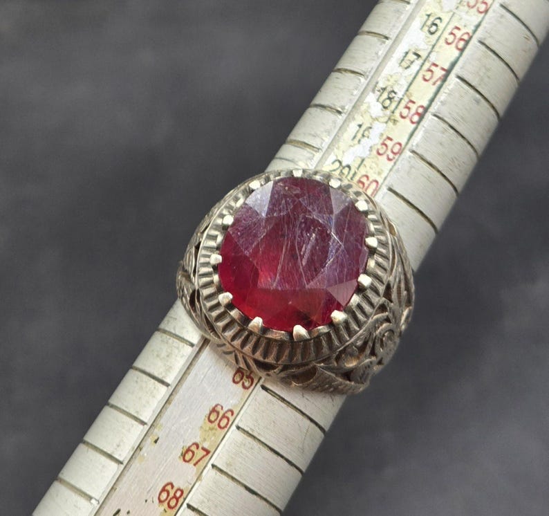 May include: A silver ring with an ornate design and a large, red gemstone. The ring is being measured with a ruler, and the measurement is 67.