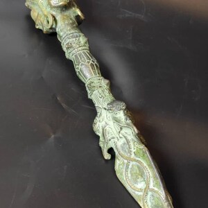 May include: A green bronze ceremonial dagger with a detailed, ornate design. The handle features a sculpted head with a fierce expression. The blade is pointed and has a decorative pattern.