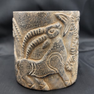 May include: A black stone cup featuring a detailed carving of an animal, possibly a goat or antelope, with intricate horns and a textured surface.