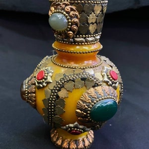 Op de afbeelding: Een gele keramische vaas met ingewikkelde versieringen van goud en zilver, waaronder kleine edelstenen in tinten groen en rood.