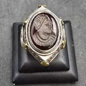 Puede incluir: Un anillo de plata con una piedra de cornalina roja que presenta un retrato tallado de una persona con casco. El anillo está decorado con detalles dorados.