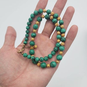 May include: A green malachite and gold bead necklace. The necklace is made of round beads with a green and white marbled pattern. The beads are separated by gold beads with a textured surface.