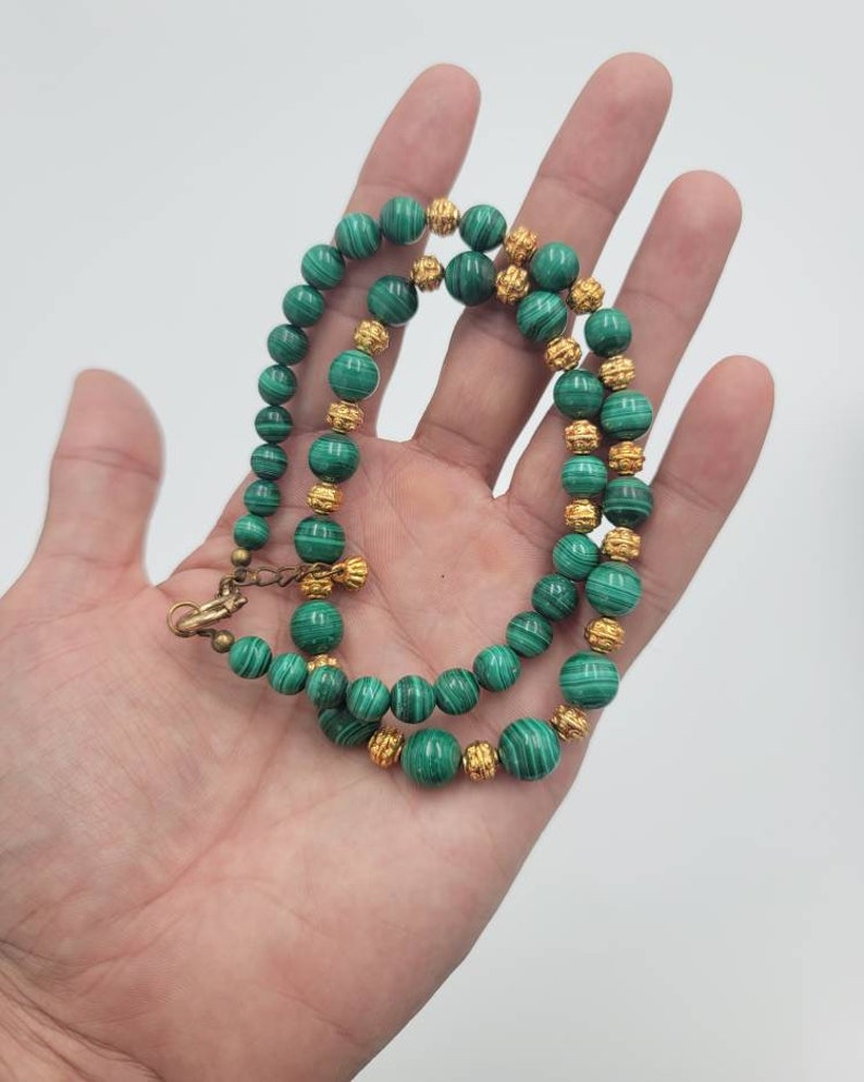 May include: A green malachite and gold bead necklace. The necklace is made of round beads with a green and white marbled pattern. The beads are separated by gold beads with a textured surface.