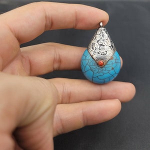 May include: A turquoise pendant with a silver filigree design and a small red stone accent. The pendant is shaped like a teardrop and has a cracked turquoise surface.