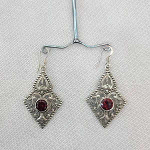May include: A pair of silver dangle earrings with a red gemstone in the center of each. The earrings have a delicate, intricate design.