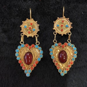 Può includere: Un paio di orecchini d'oro con pietre rosse e blu. Gli orecchini hanno la forma di un cuore e un ciondolo rotondo sopra il cuore.