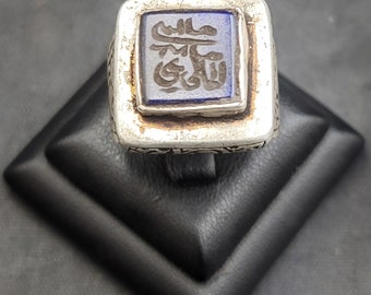 Può includere: Un anello d'argento con una pietra quadrata blu incastonata in una cornice d'argento. La pietra è incisa con caratteri arabi.