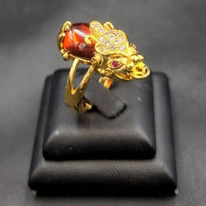 May include: A gold ring with a red gemstone and a detailed dragon design. The dragon's eyes are set with red stones and the rest of the ring is adorned with clear stones.