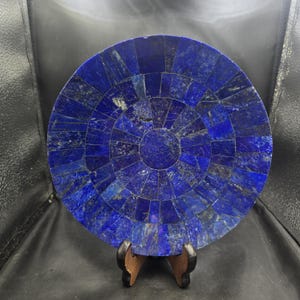 Puede incluir: Un plato redondo de mosaico de lapislázuli azul sobre un soporte de madera. El plato está hecho de muchas piezas pequeñas e irregulares de lapislázuli, creando un patrón hermoso e intrincado.