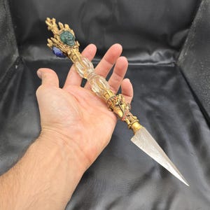 May include: An ornate, gold-colored ritual dagger with a clear, faceted handle and a sharp, triangular blade. The hilt features intricate carvings and is adorned with green and blue gemstones. The dagger is held in a hand against a dark background.