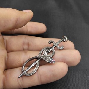 Può includere: Un ciondolo d'argento a forma di serpente avvolto attorno a una spada. Il serpente e la spada sono entrambi ricoperti di piccole pietre trasparenti. Gli occhi del serpente sono rossi.