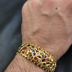 May include: A gold bangle bracelet with a multi-colored gemstone design. The bracelet is adorned with a variety of round, square, and pear-shaped gemstones in shades of red, pink, purple, green, and yellow.