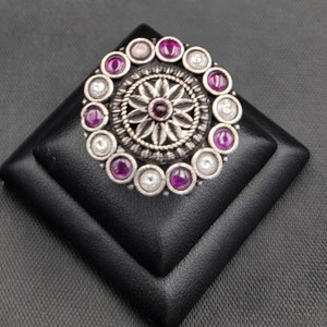 Może przedstawiać: Srebrny pierścionek z okrągłym wzorem, w którym znajduje się centralny kwiat z fioletowym kamieniem otoczonym kółkiem z naprzemiennie fioletowych i białych kamieni.